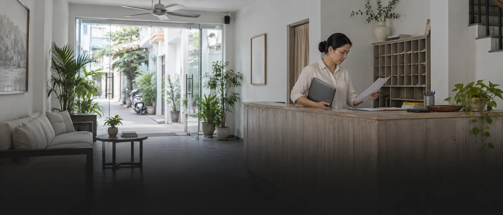 Chủ một khách sạn nhỏ đứng sau quầy lễ tân buổi sáng sớm, một tay kẹp máy tính xách tay đã gập, tay kia cầm bảng giá in.