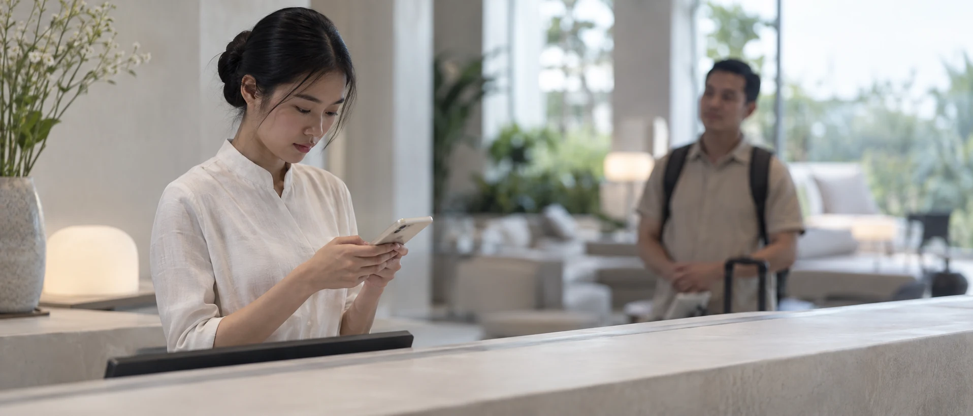 Nhân viên lễ tân đứng sau quầy soạn tin trả lời trên điện thoại, phía sau có một người khách đang chờ.
