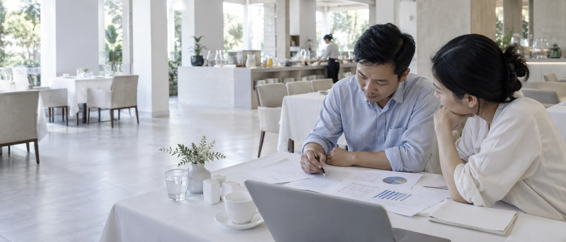 Hai người ngồi cùng một phía bàn trong phòng ăn sáng đã vãn khách, các trang in trải trước mặt.